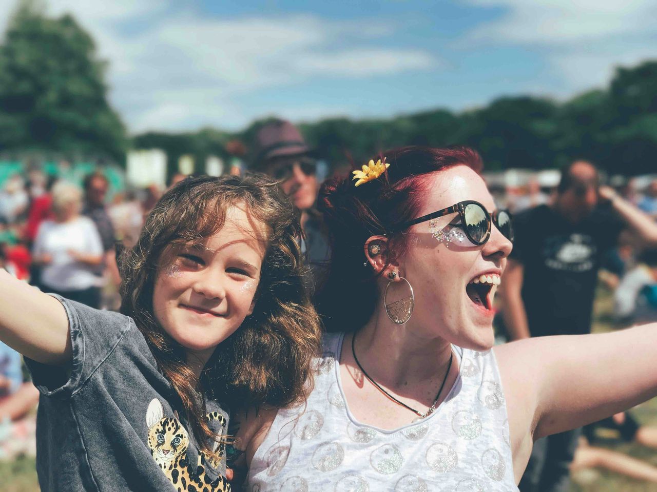 Dos amigas (mujer joven pelirroja con lentes de sol y nena castaña) abrazadas riendo con brazos abiertos en festival al aire libre, ejemplo de fotografía lifestyle natural