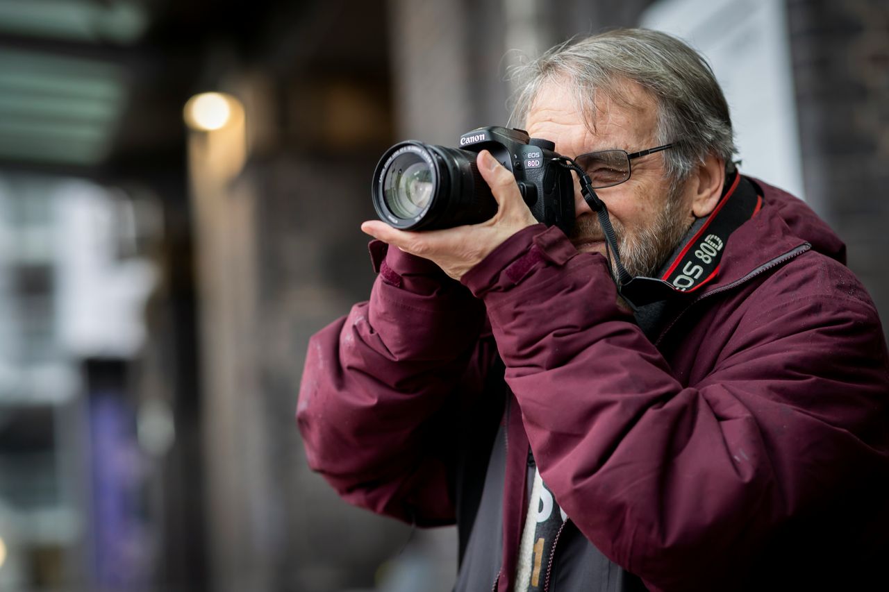 Hombre mayor con lentes y campera bordo sacando foto con Canon DSLR en la calle, ejemplo de fotografía documental urbana
