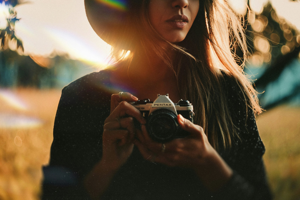 Mujer con sombrero sosteniendo cámara vintage al atardecer en campo dorado, composición con regla de tercios, contraluz y lens flare, ejemplo de composición fotográfica artística