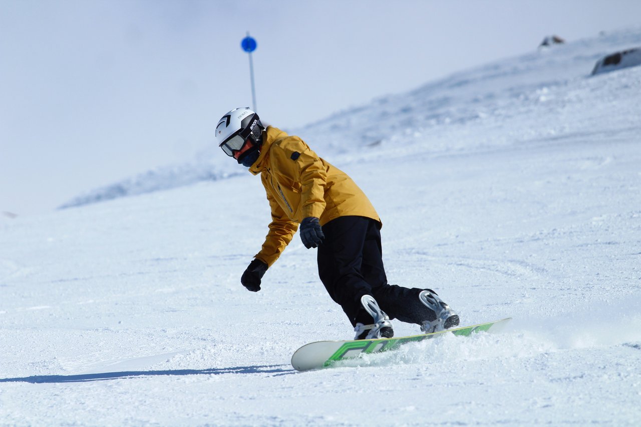 Snowboarder con campera amarilla y casco blanco descendiendo pista nevada del Cerro Catedral en día soleado, ejemplo de fotografía deportiva en alta montaña argentina