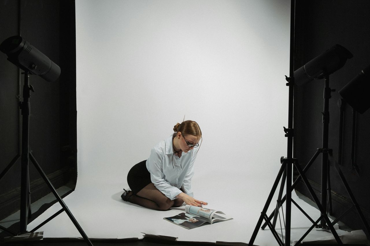 Fotógrafa con lentes sentada en piso de estudio profesional rodeada de flashes y trípodes hojeando revista entre tomas, reflejo de las largas jornadas de trabajo en sesiones fotográficas