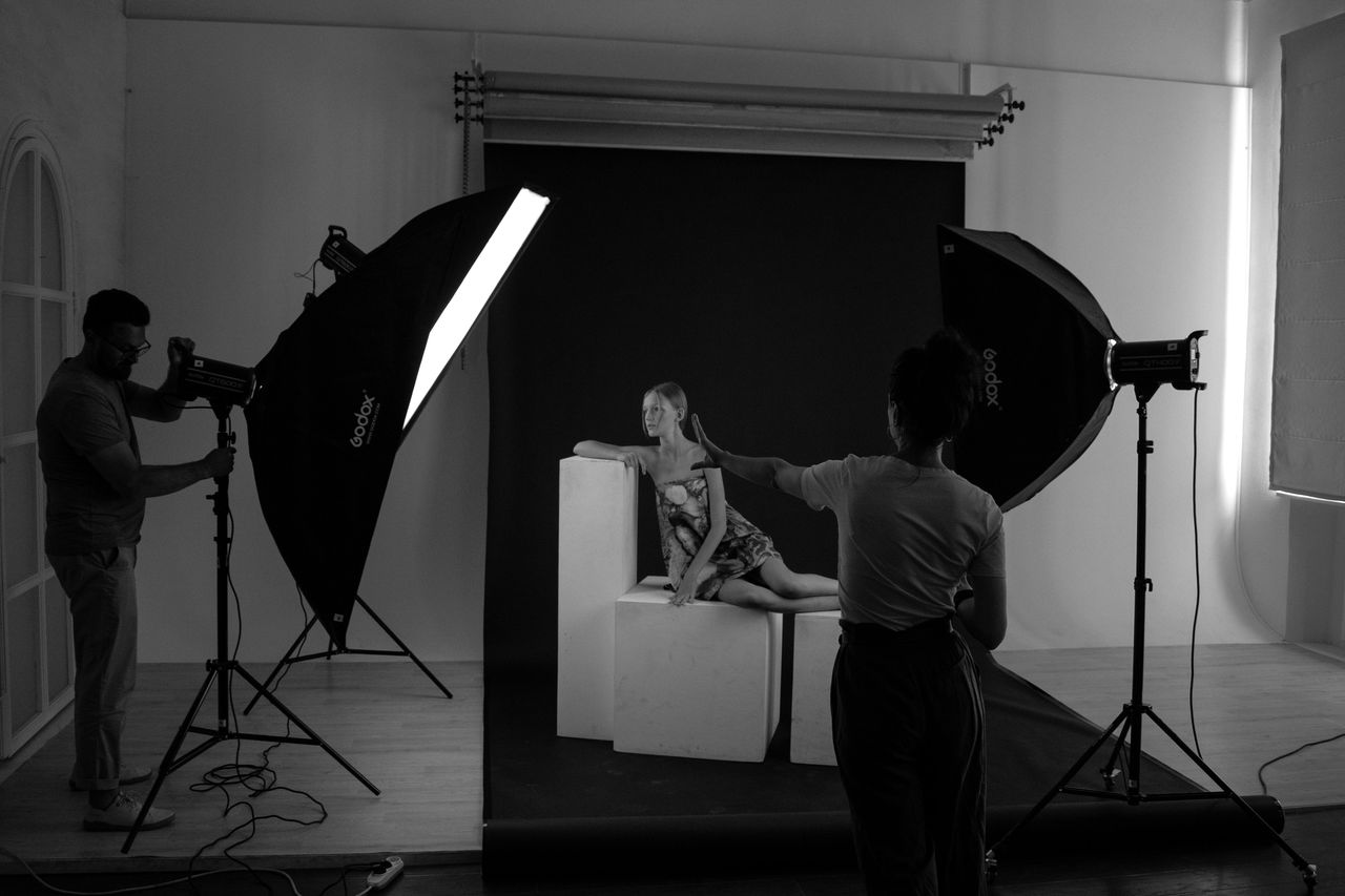 Asistente de fotografía ajustando softbox Godox en estudio profesional con fondo negro mientras el fotógrafo dirige al modelo sentado en cubo, sesión de fotos en blanco y negro