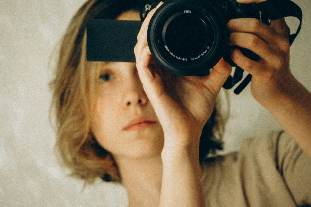 Mujer joven rubia de pelo corto sosteniendo cámara Leica vintage frente a su rostro, recostada con remera beige, tono cálido nostálgico, ejemplo de fotografía con estética vintage