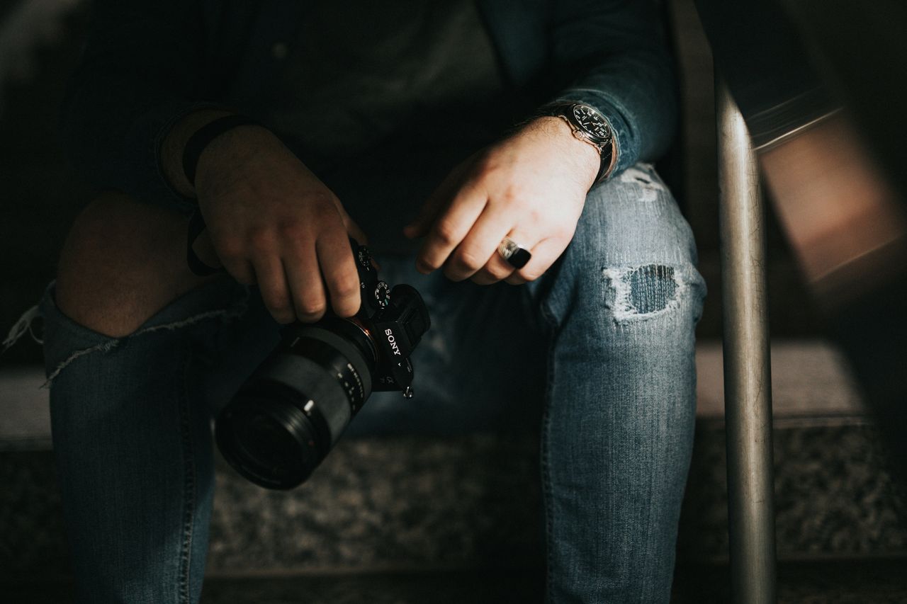 Manos masculinas con jeans rotos, anillo negro y reloj sosteniendo cámara Sony mirrorless sobre escalones, tono moody oscuro cinematográfico, simbolizando equipo profesional con C2PA
