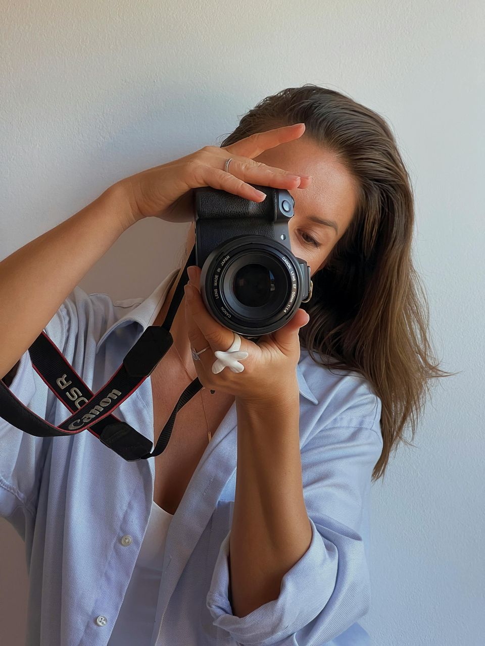 Mujer joven con camisa celeste oversize sosteniendo una Canon DSLR contra fondo claro minimalista, ejemplo de fotografía minimalista con espacio negativo