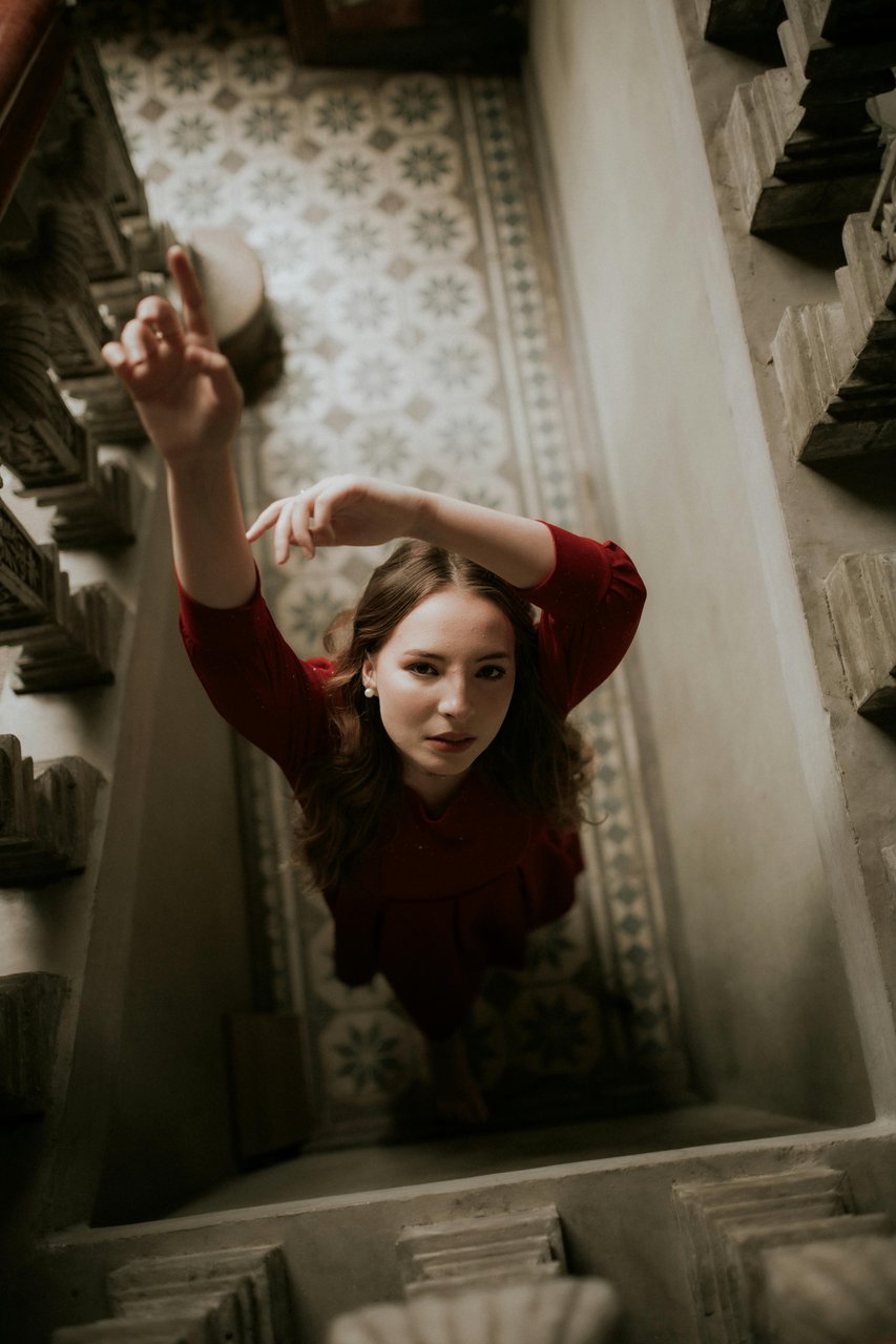 Retrato editorial de joven con vestido rojo extendiendo los brazos hacia arriba, vista en picado desde una escalera con piso de mosaico floral, sesión de producción de quince años