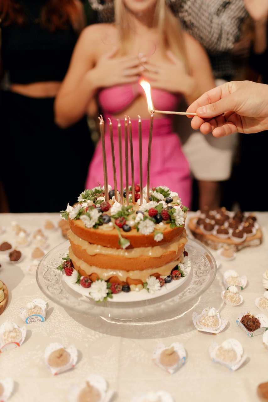 Mano encendiendo velas finas sobre una torta naked decorada con frutos rojos y flores en la mesa dulce de una fiesta, luz tenue de salón