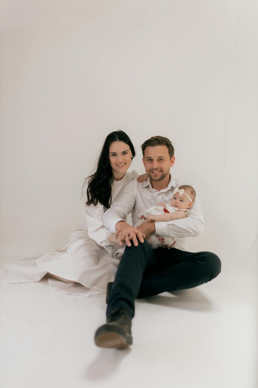 Papá y mamá sentados sobre un fondo blanco de estudio con su bebé en brazos, vestidos en tonos blancos y negros, durante una sesión de fotos de recién nacido con papás