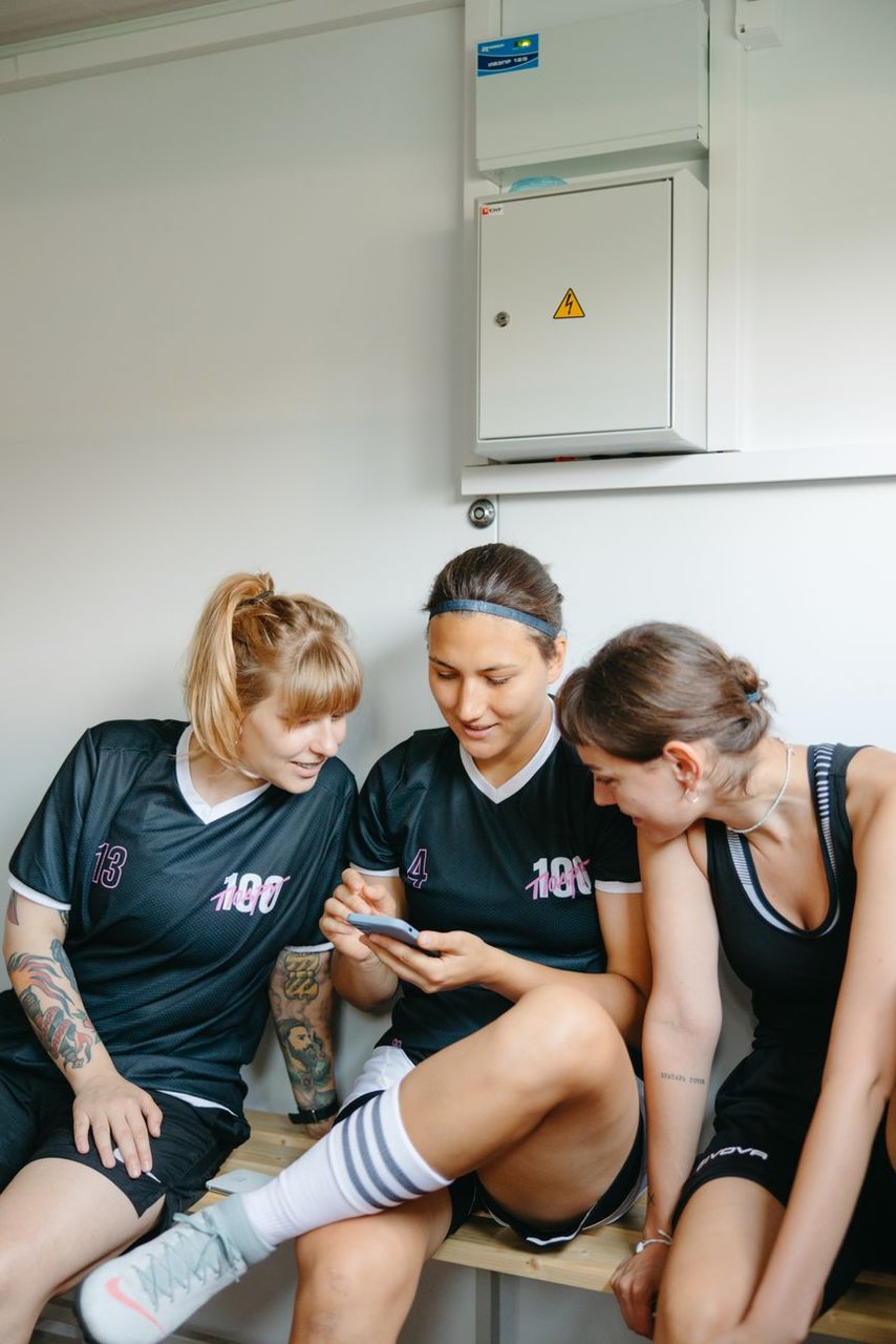 Tres jugadoras jóvenes con camisetas deportivas numeradas sentadas en un banco de vestuario mirando juntas la pantalla de un celular
