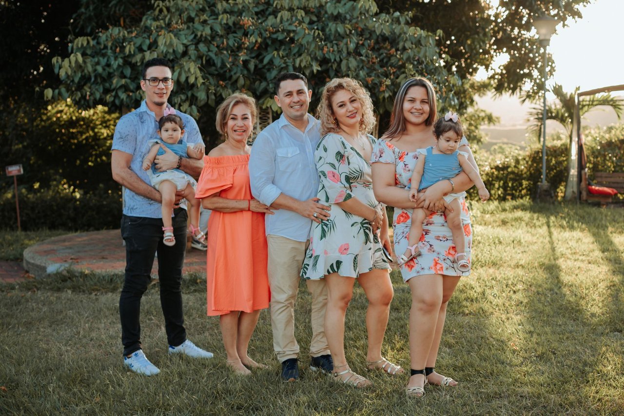 Familia numerosa de tres generaciones posando en el jardín de una casa al atardecer, con dos bebés en brazos