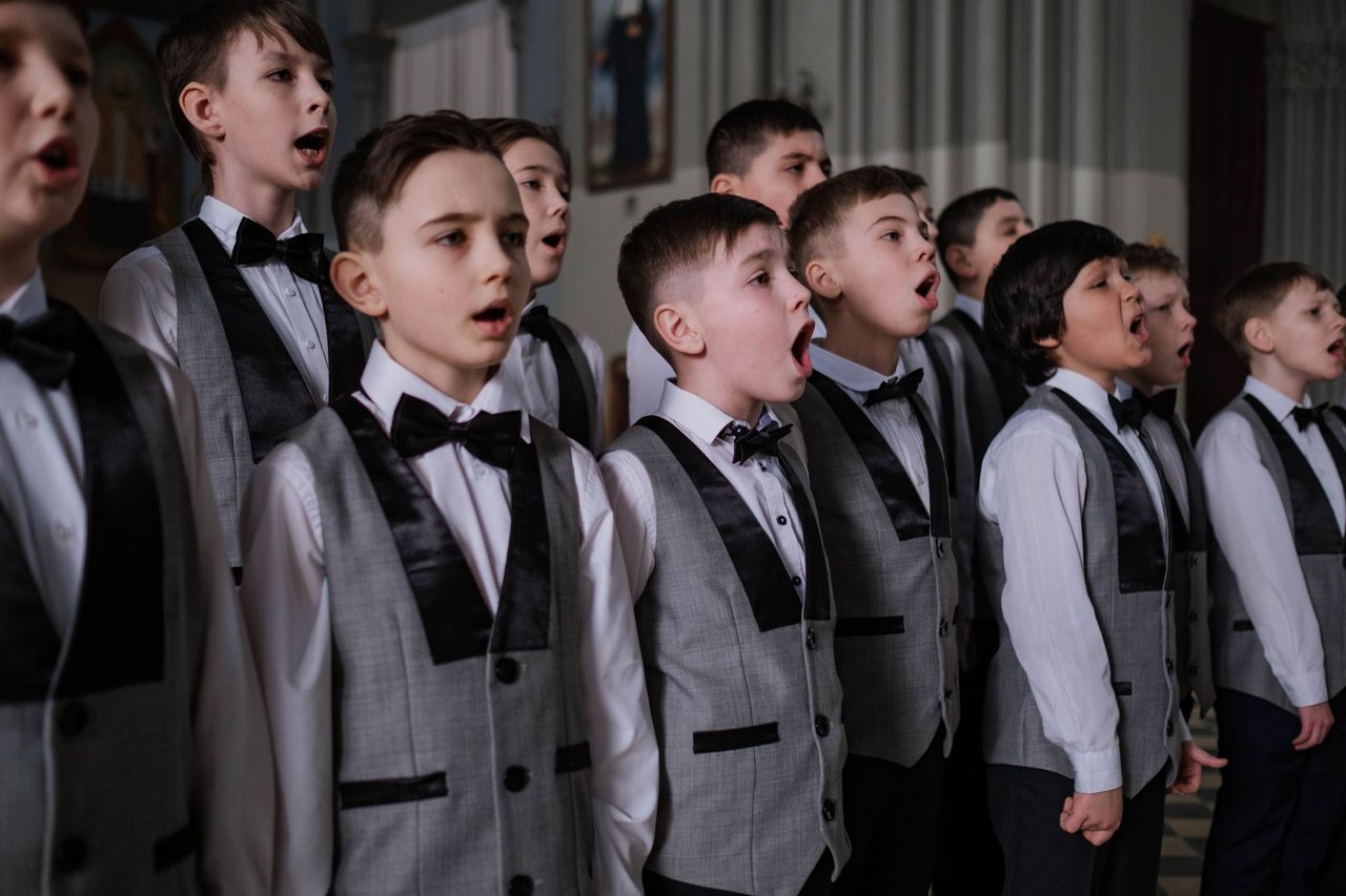 Coro de chicos con chaleco y moño cantando durante un acto escolar en un salón con luz tenue, la situación típica de poca luz en el SUM