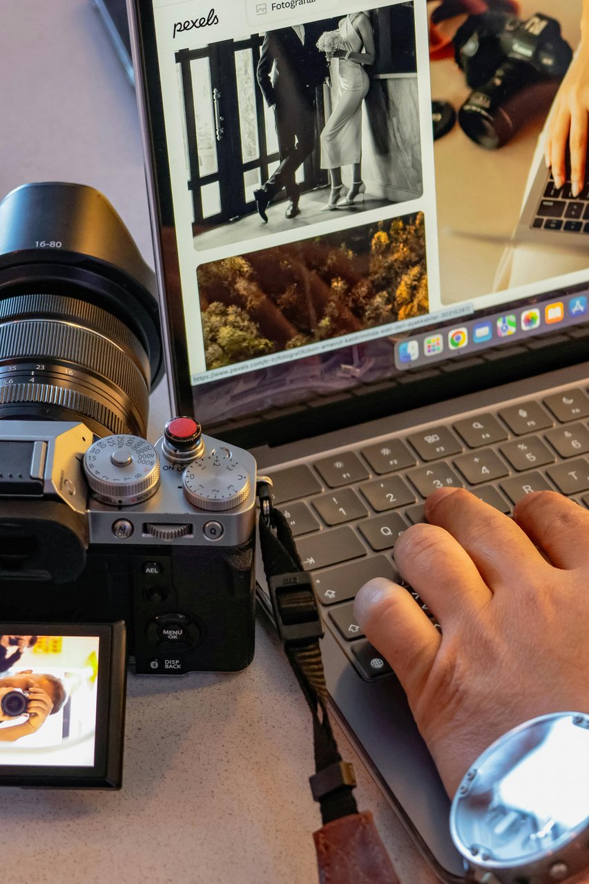 Escritorio con cámara Fujifilm mirrorless y notebook mostrando un sitio de banco de imágenes con fotos en grilla, manos tipeando en el teclado
