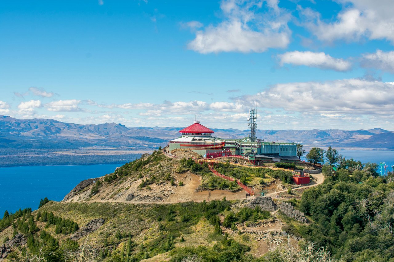 Cima del Cerro Otto con su confitería giratoria de techo rojo y antena, vista al Nahuel Huapi y montañas patagónicas de fondo en día soleado