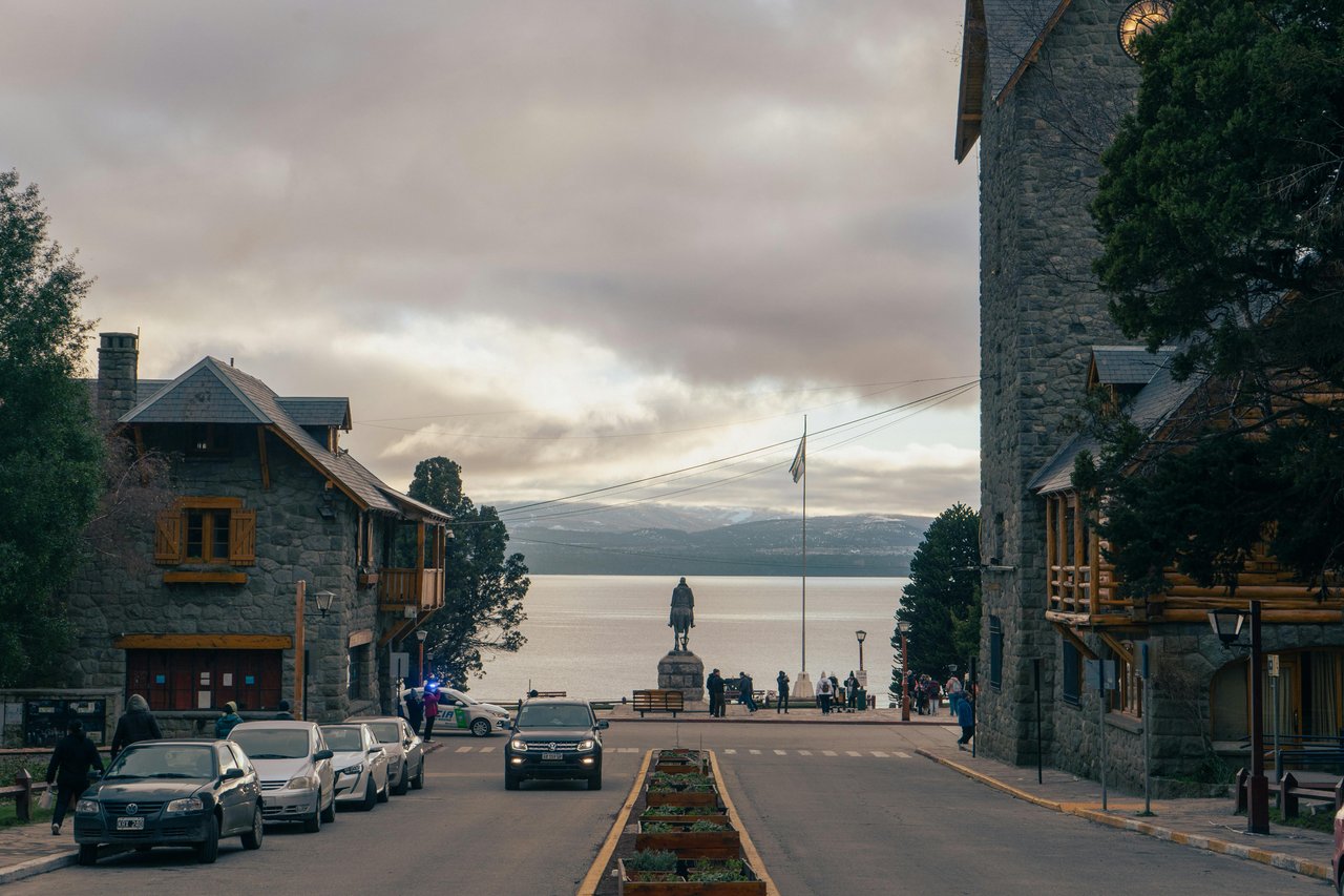 Calle del Centro Cívico de Bariloche con edificios de piedra y techos a dos aguas, vista al Nahuel Huapi al fondo con monumento ecuestre central, atmósfera invernal patagónica