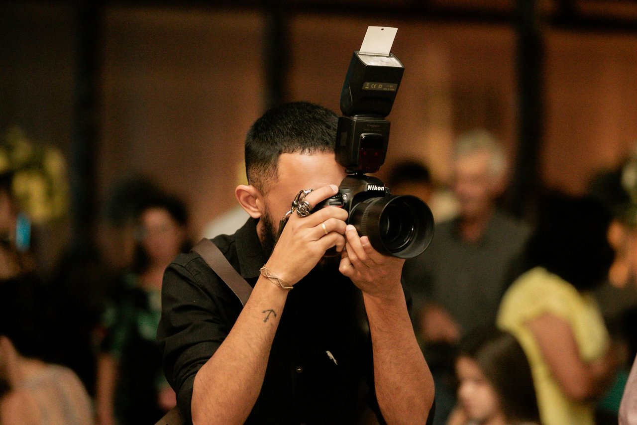 Fotógrafo con cámara Nikon y flash externo enfocando en un evento social con multitud desenfocada y luces cálidas de fondo, cobertura nocturna de salón