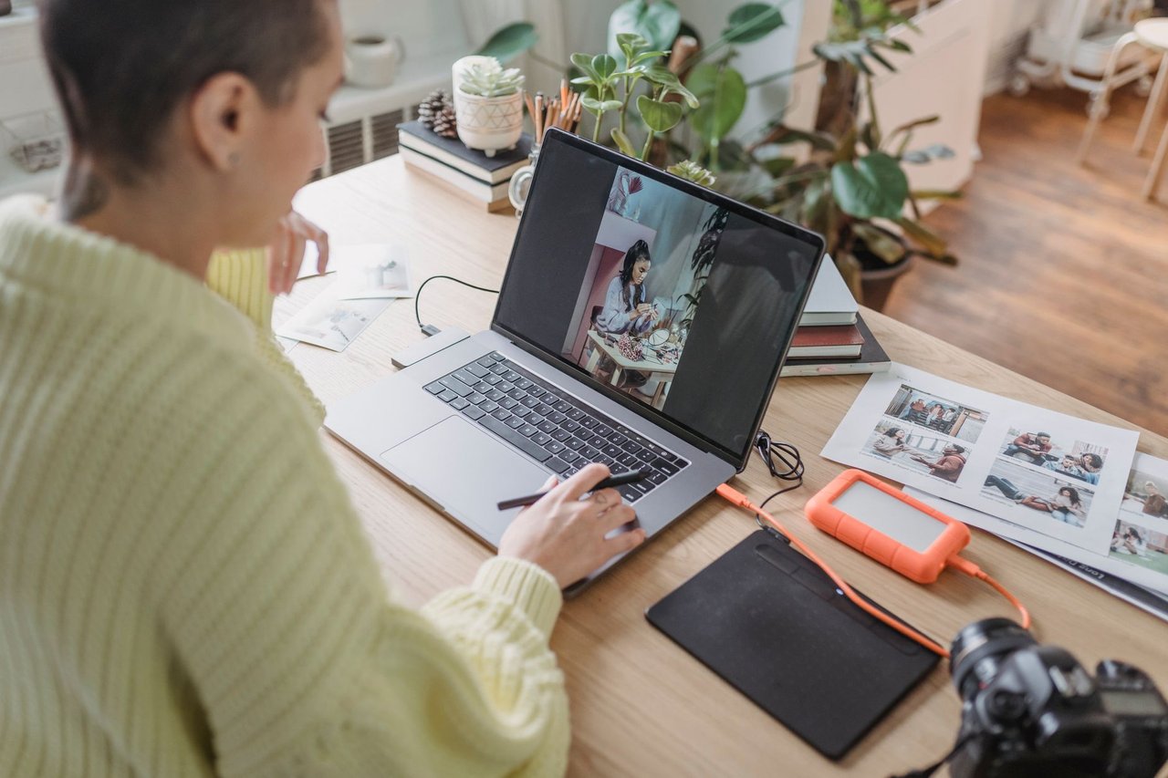 Fotógrafa editando fotos en una laptop con tableta gráfica y copias impresas sobre el escritorio, comparando la pantalla contra el papel