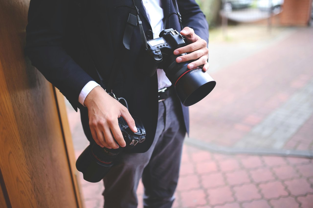 Fotógrafo con traje oscuro sosteniendo dos cuerpos de cámara Canon con teleobjetivos en un pasillo de ladrillos, listo para cubrir un casamiento con equipo de respaldo