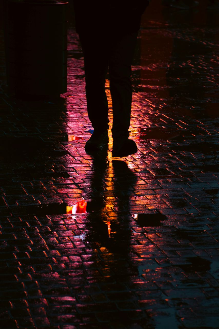 Silueta de persona caminando en adoquines mojados con reflejos rosas y naranjas de neón, escena nocturna urbana, ejemplo de fotografía con estilo cinematic