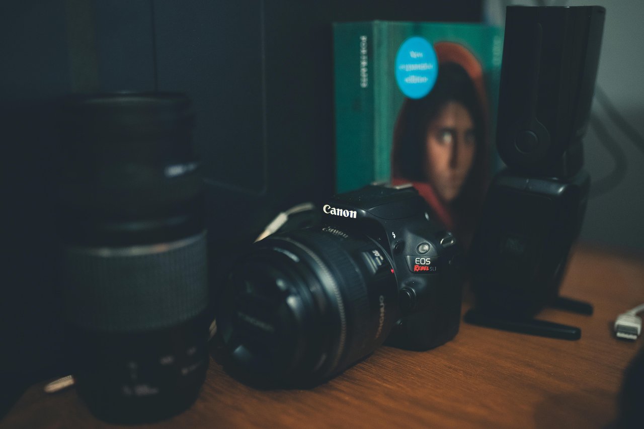 Cámara Canon EOS Rebel de entrada con su lente apoyada sobre un escritorio de madera junto a un flash externo y un libro de fotografía, ejemplo de equipo accesible para empezar