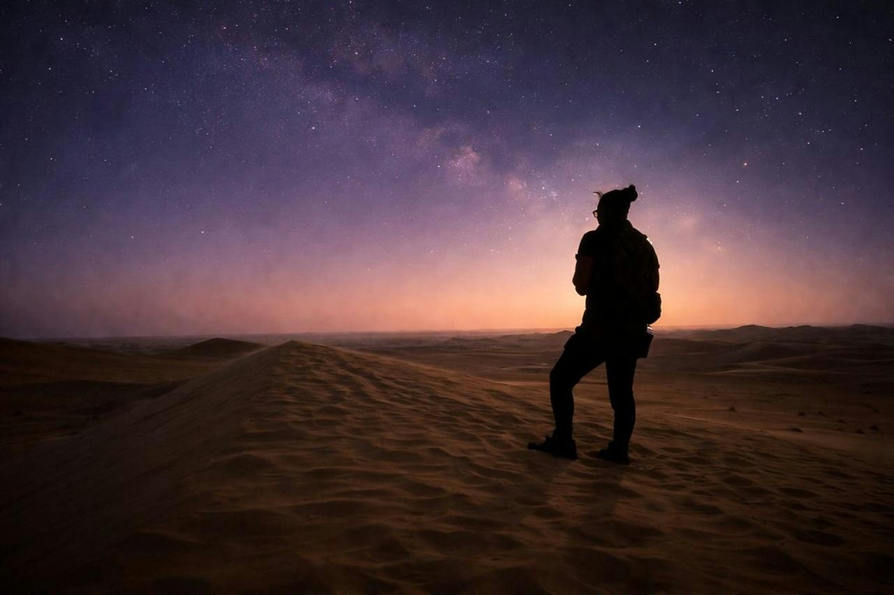 Silueta de una persona con mochila parada sobre una duna bajo el arco de la Vía Láctea, el primer plano que le da escala y lugar a la foto