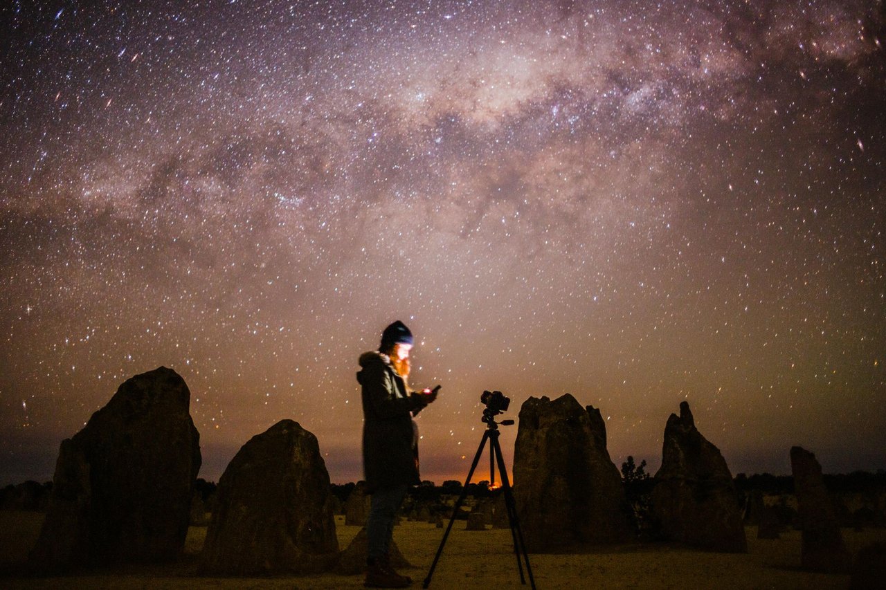 Fotógrafo parado junto a su cámara montada en trípode en un desierto de formaciones rocosas, con la Vía Láctea sobre el horizonte y una linterna encendida en la mano
