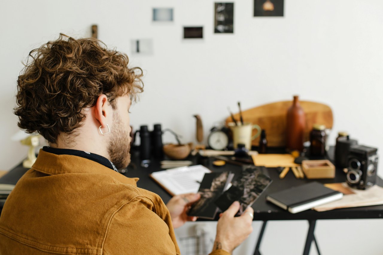 Fotógrafo de espaldas revisando copias impresas sobre un escritorio negro junto a una cámara TLR vintage, controlando el resultado de la impresión