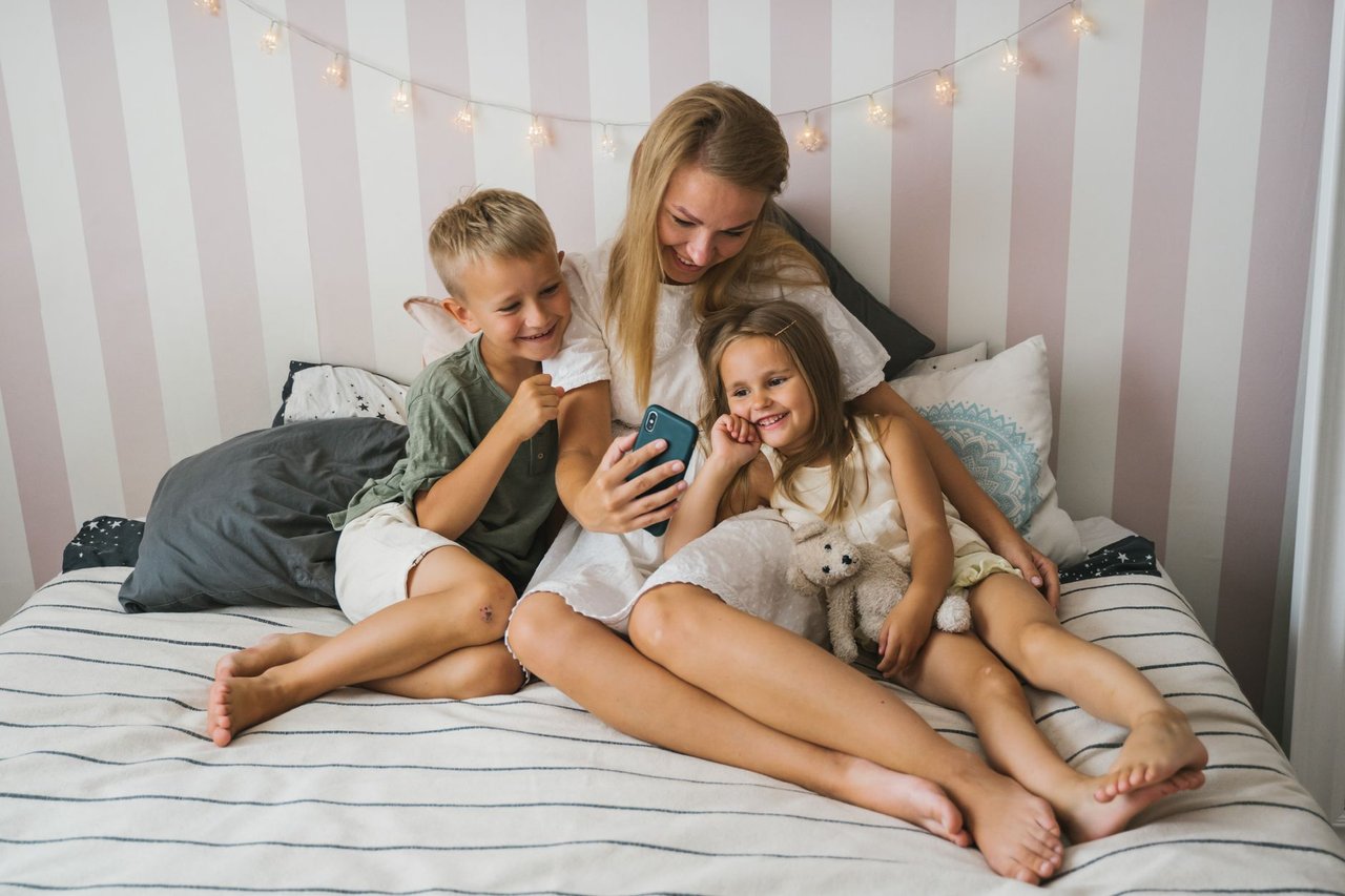 Madre y dos hijos sentados en la cama mirando juntos la pantalla de un celular, la familia accede a las fotos del curso desde su casa