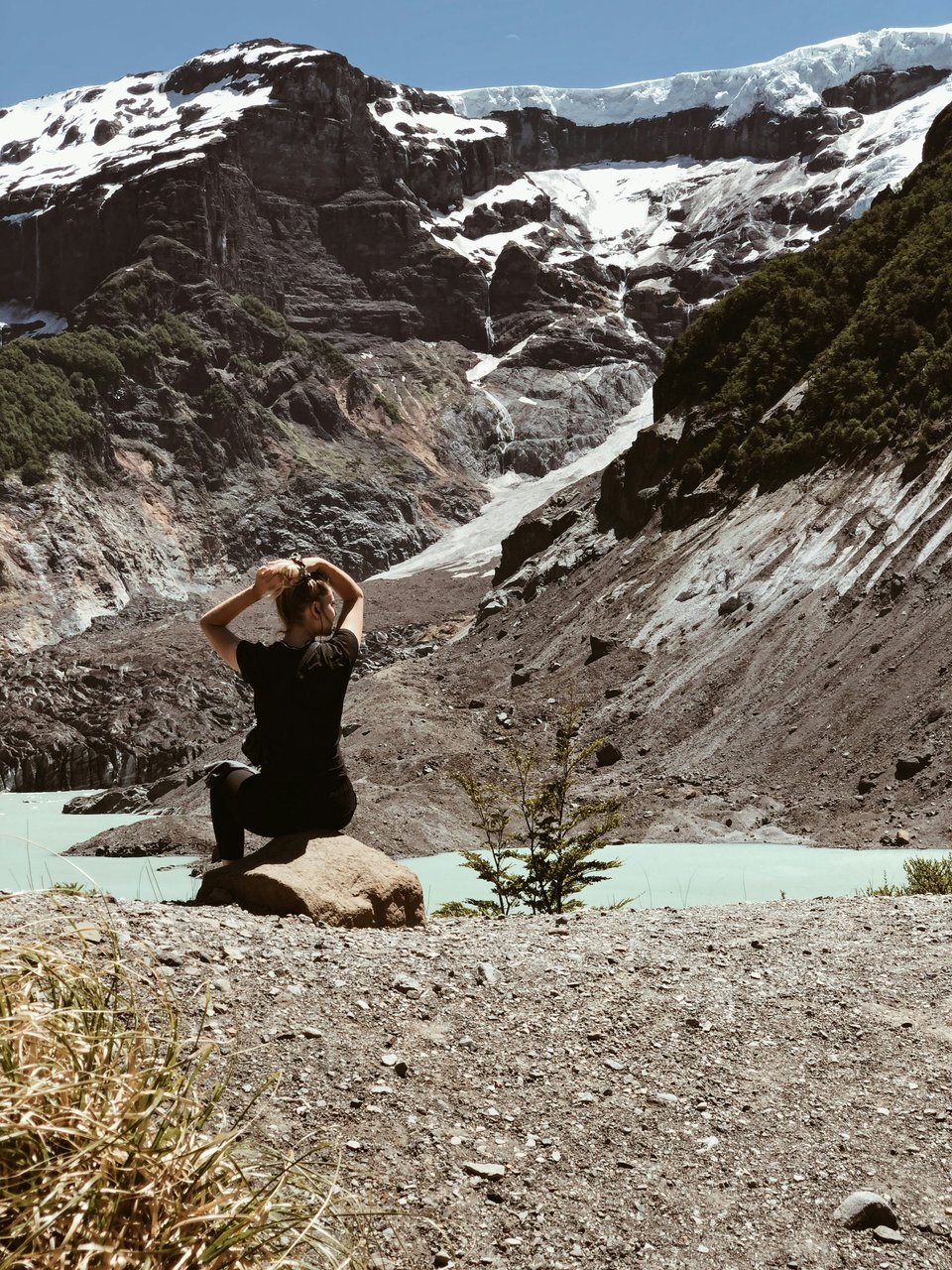 Mujer joven sentada en piedra frente al Ventisquero Negro del Cerro Tronador, glaciar colgante visible y laguna glaciaria gris al fondo, ejemplo de uso de figura humana como escala en fotografía de paisaje patagónico