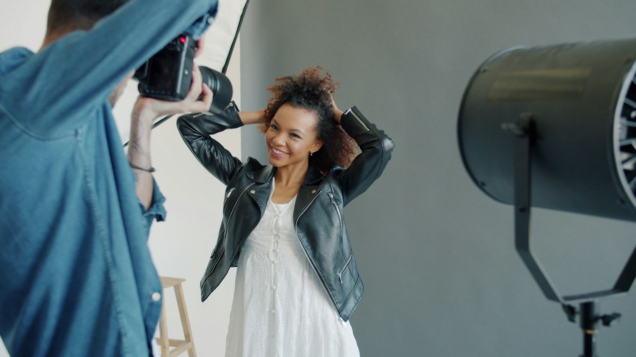 Mujer con campera de cuero y pelo rizado con manos en el pelo, sonrisa amplia y natural durante sesión de fotos en estudio