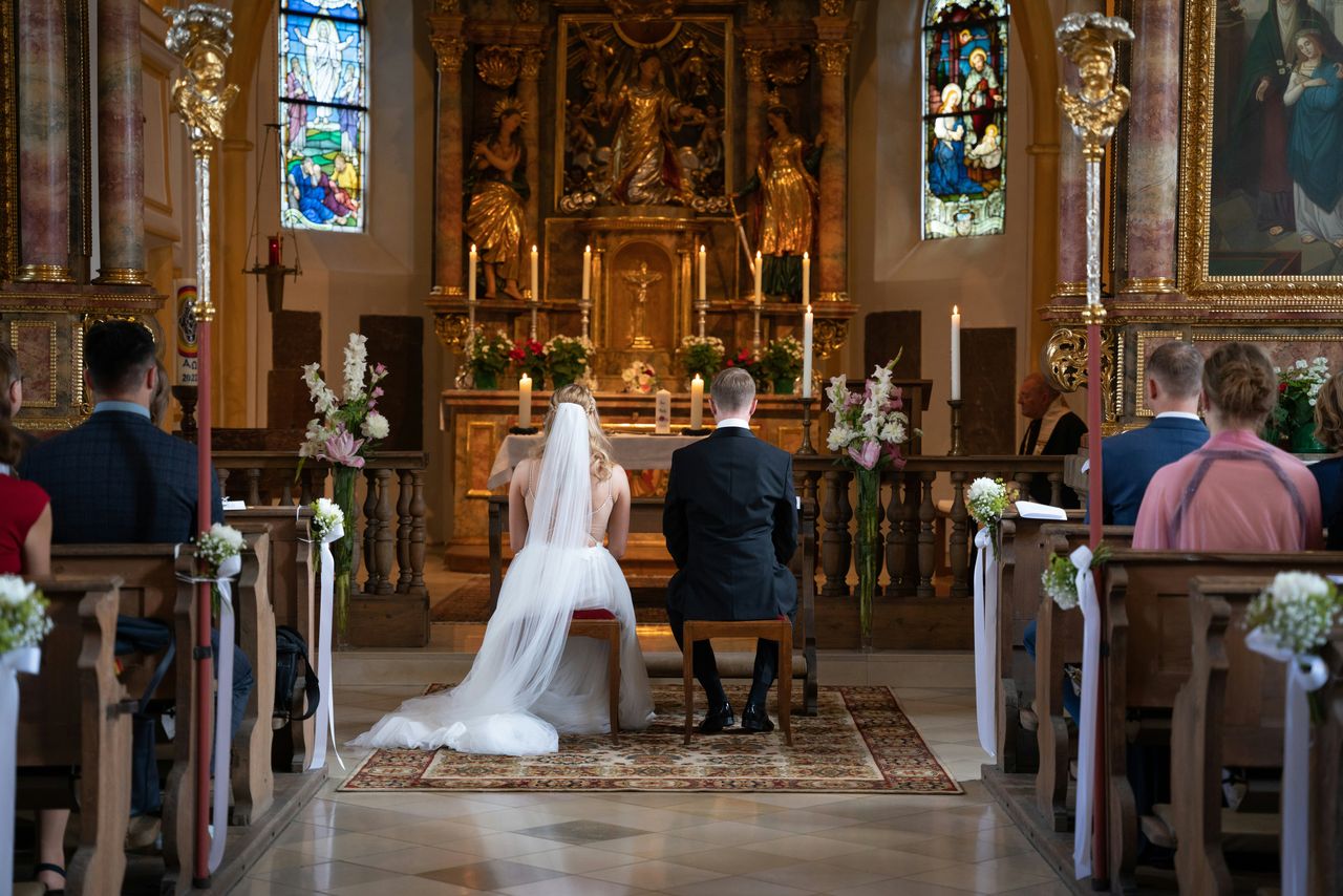 Pareja de novios arrodillados frente al altar dorado de una iglesia barroca durante la ceremonia, con vitrales iluminados y composición simétrica, escena de poca luz típica del casamiento